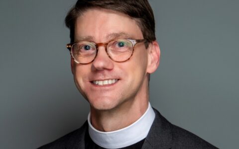 Kevin Sparrow, A white man with white priest's collar, glasses, and grey jacket smiles at camera.
